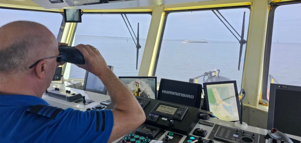 Een Rijkswaterstaat medewerker kijkt met zijn verrekijker over het water, vanaf een patrouillevaartuig.